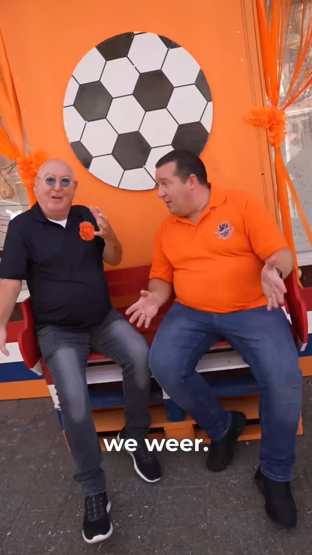 Stef Ekkel en Harry Vermeegen in oranje op een bankje, met een grote voetbal op de achtergrond